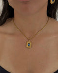 A close-up of a woman wearing a gold chain necklace with a rectangular gold pendant featuring a blue center stone and textured border, along with matching gold hoop earrings.