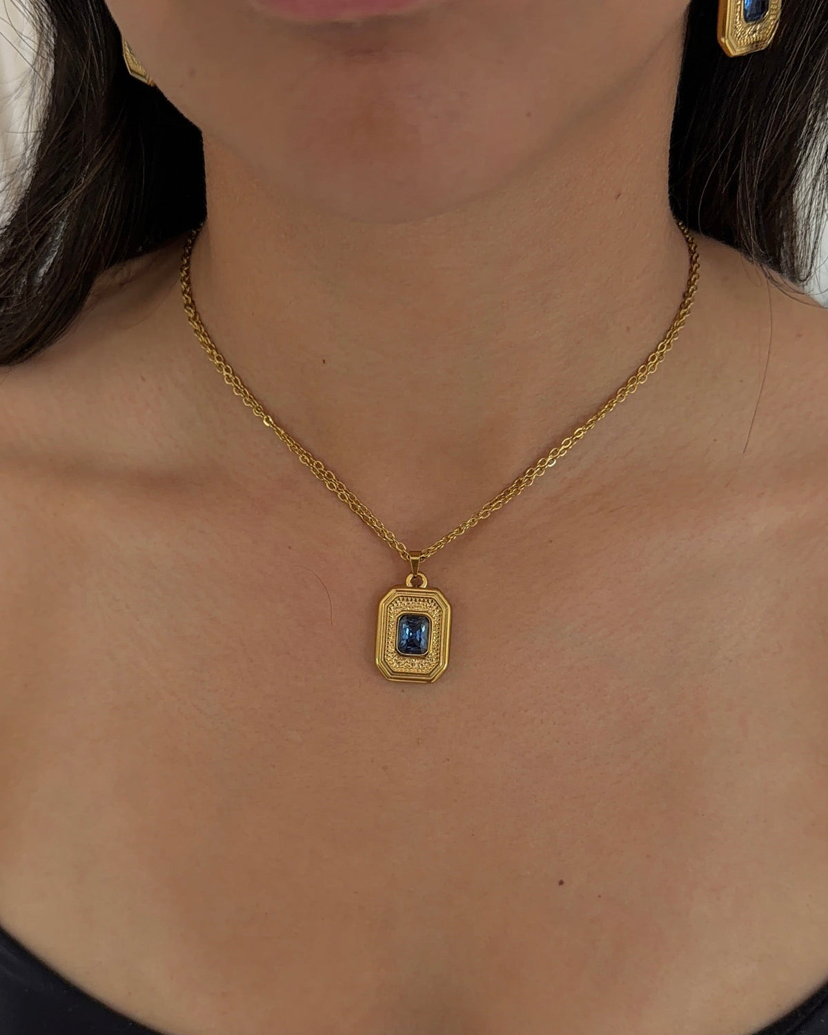 A close-up of a woman wearing a gold chain necklace with a rectangular gold pendant featuring a blue center stone and textured border, along with matching gold hoop earrings.