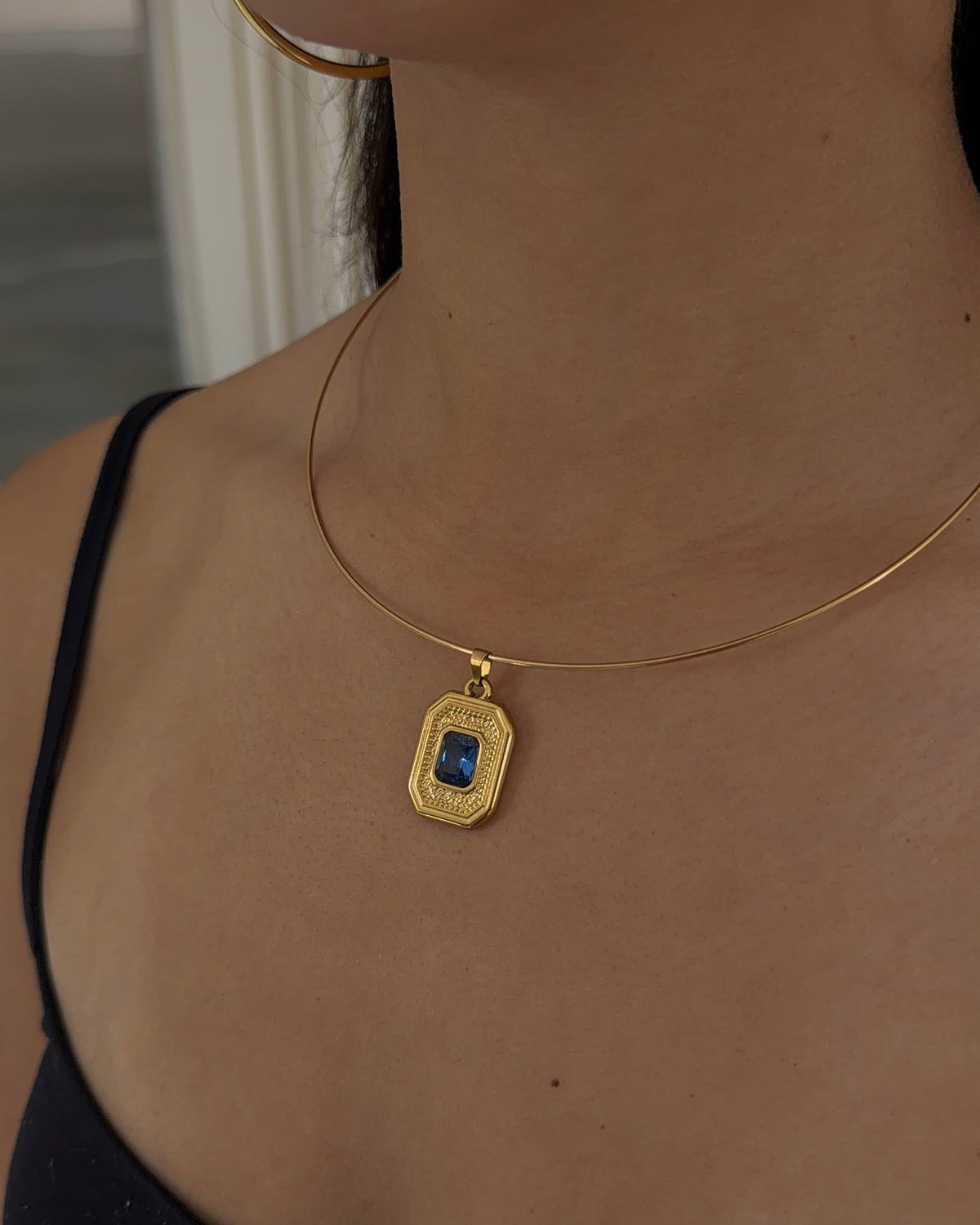 A close-up of a model wearing a delicate gold chain necklace with a rectangular gold pendant featuring a blue stone in the center.