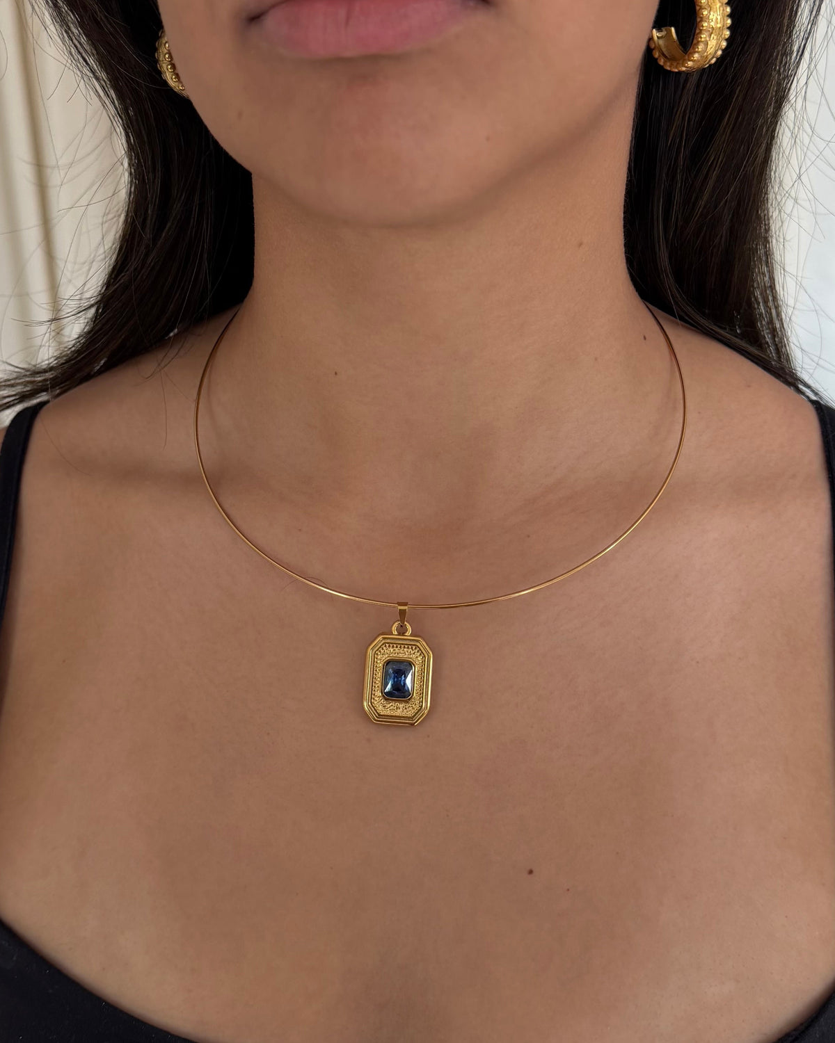 A woman wearing a delicate gold necklace with a rectangular pendant featuring a blue and white pattern, paired with gold hoop earrings and a black spaghetti-strap top.
