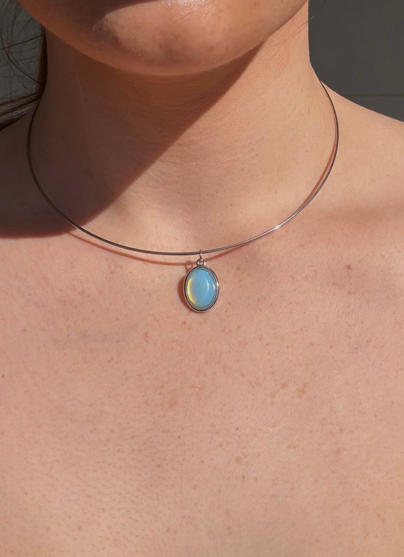 A close-up of a person wearing a delicate silver-toned necklace with a small oval light blue pendant, shown outdoors against a light gray wall.