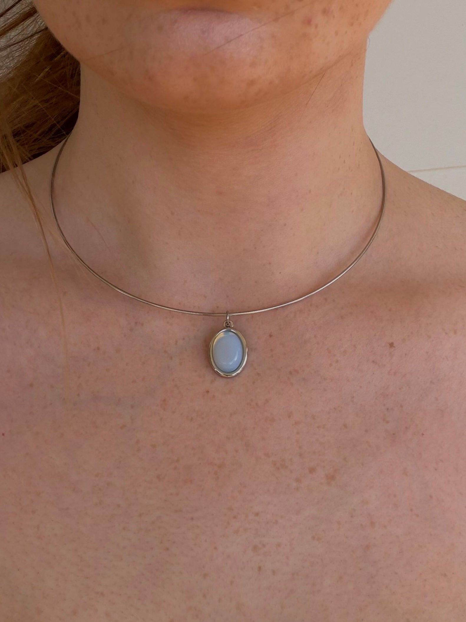 A close-up of a person's neck and chest wearing a delicate silver wire choker necklace with a small oval light blue-gray pendant.