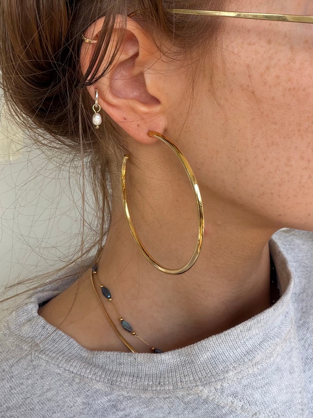 A close-up of a person wearing a large polished gold hoop earring, with additional delicate gold jewelry including a small pearl drop and layered necklaces visible.