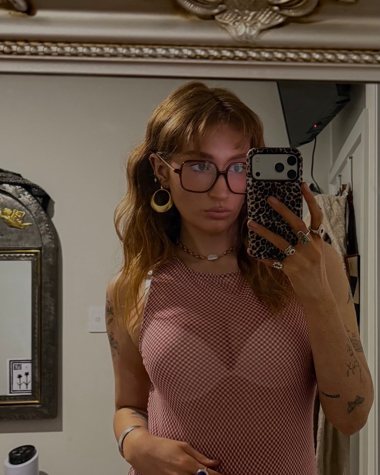 A woman taking a mirror selfie wearing a pink fishnet sleeveless top, black eyeglasses, and multiple rings, in an indoor room setting.