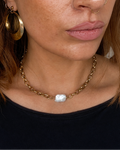A close-up of a woman wearing a gold chain choker necklace with a central pearl stone, along with gold hoop earrings and a nose ring, wearing a black top.