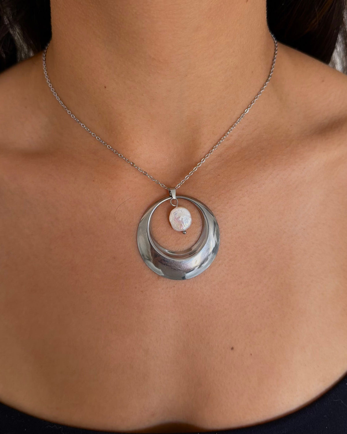 A close-up of a model wearing a silver crescent moon pendant necklace with a small white stone in the center, paired with silver hoop earrings and a black top.