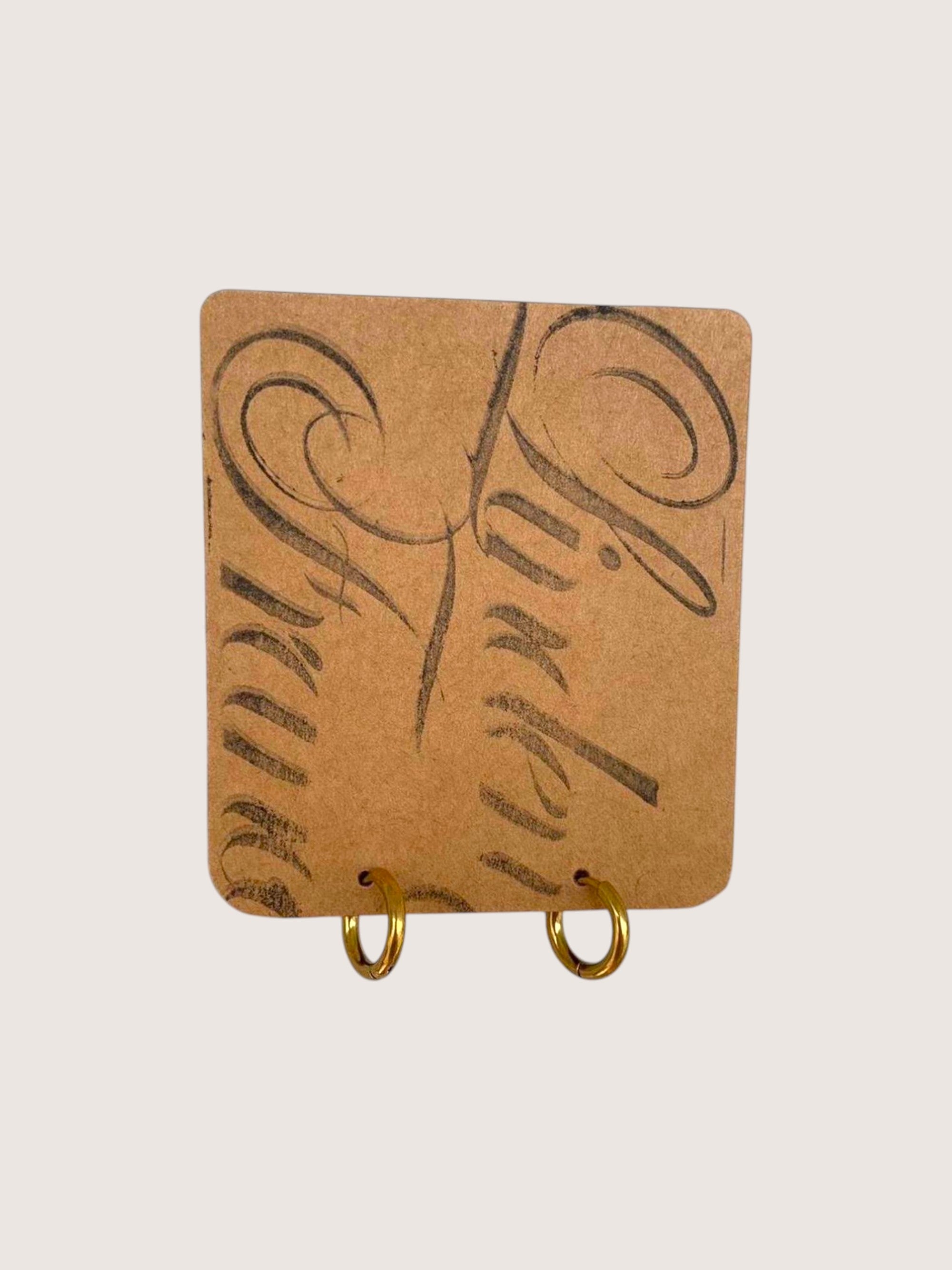 A pair of small gold hoop earrings displayed on a square tan card with abstract black markings, set against a light gray background.