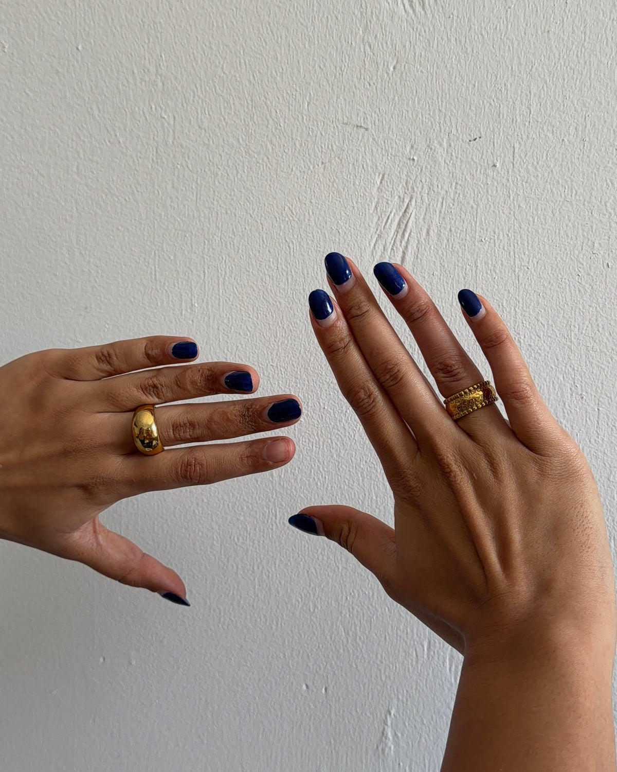 Two hands shown against a plain off-white wall, each wearing a chunky gold ring. The right hand has dark blue manicured nails, and the left hand has dark blue nail polish on the fingers and a dark blue accent on the thumb.