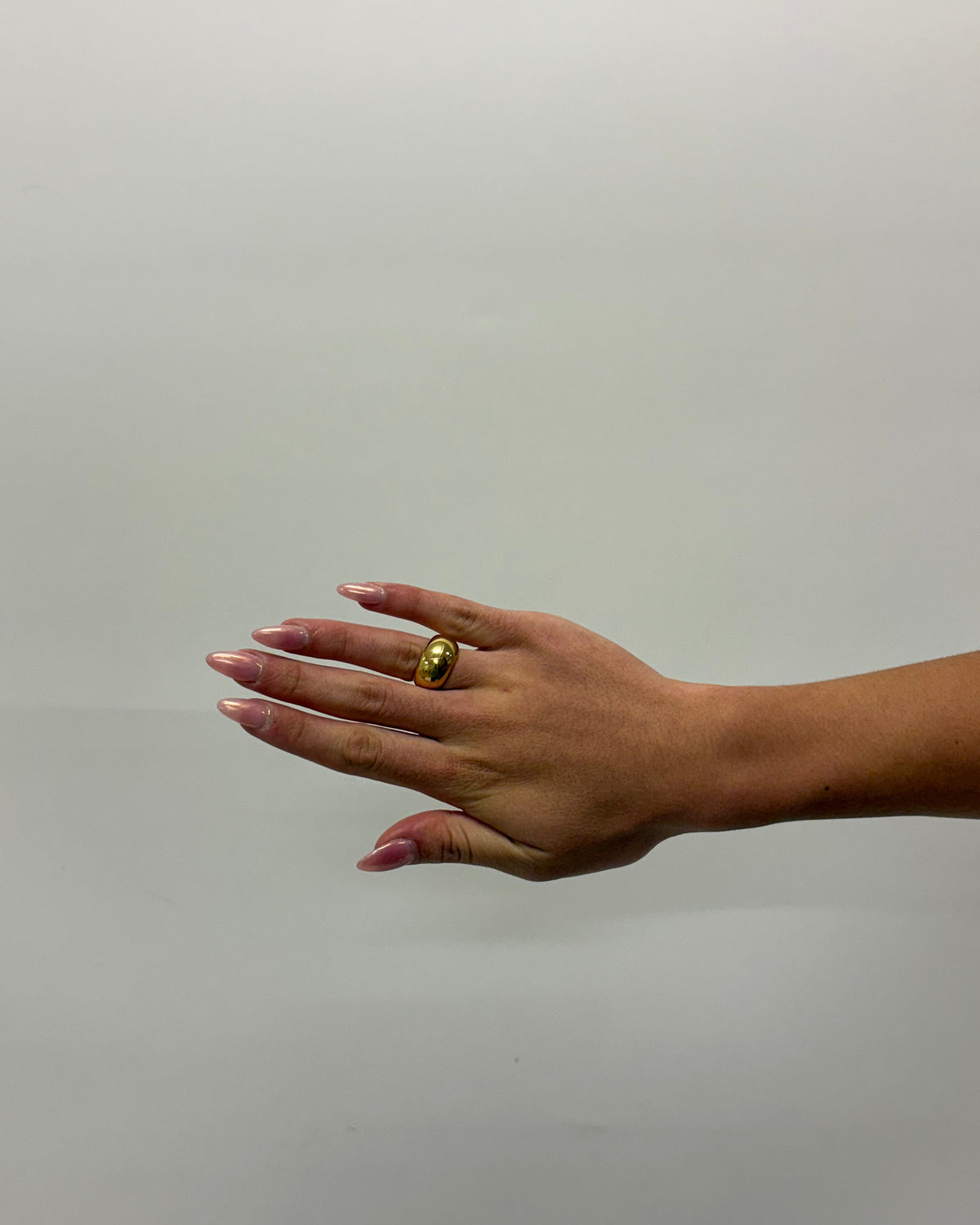 A gold oval ring with a central black stone, worn on the ring finger of a hand with pink manicured nails, set against a plain light gray background.