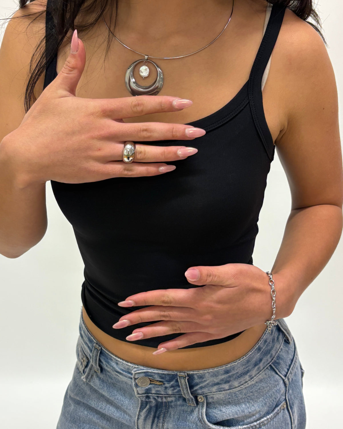 A woman wearing a black ribbed tank top and light blue denim jeans, accessorized with a silver pendant necklace, ring, and bracelet.