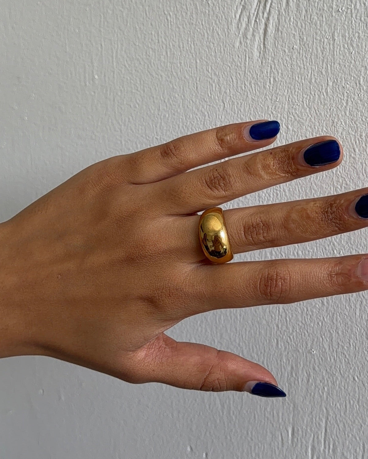A close-up of a hand wearing a chunky, polished gold ring on the ring finger, set against a neutral textured light gray background.