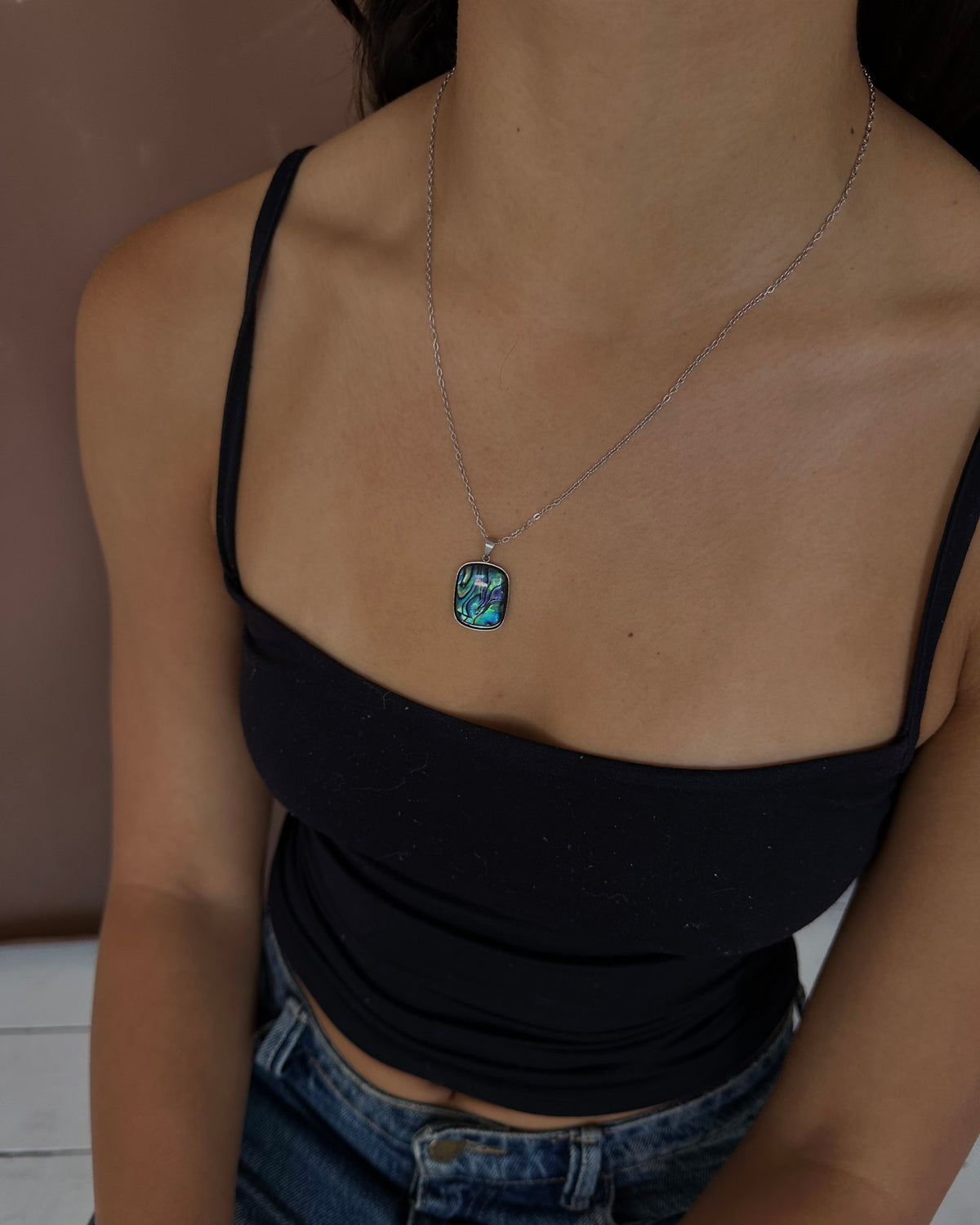 A silver-toned necklace with a rectangular turquoise-blue pendant is worn by a model in a black camisole top and blue jeans.