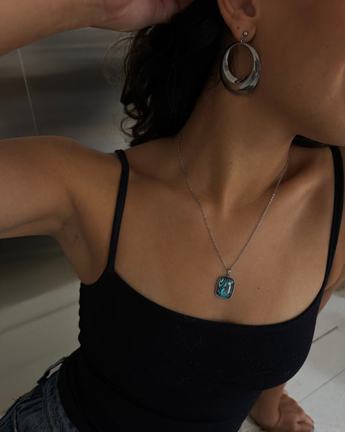 A close-up lifestyle shot of a person wearing a silver-toned necklace with a rectangular turquoise-blue pendant, paired with a black camisole top and denim shorts.