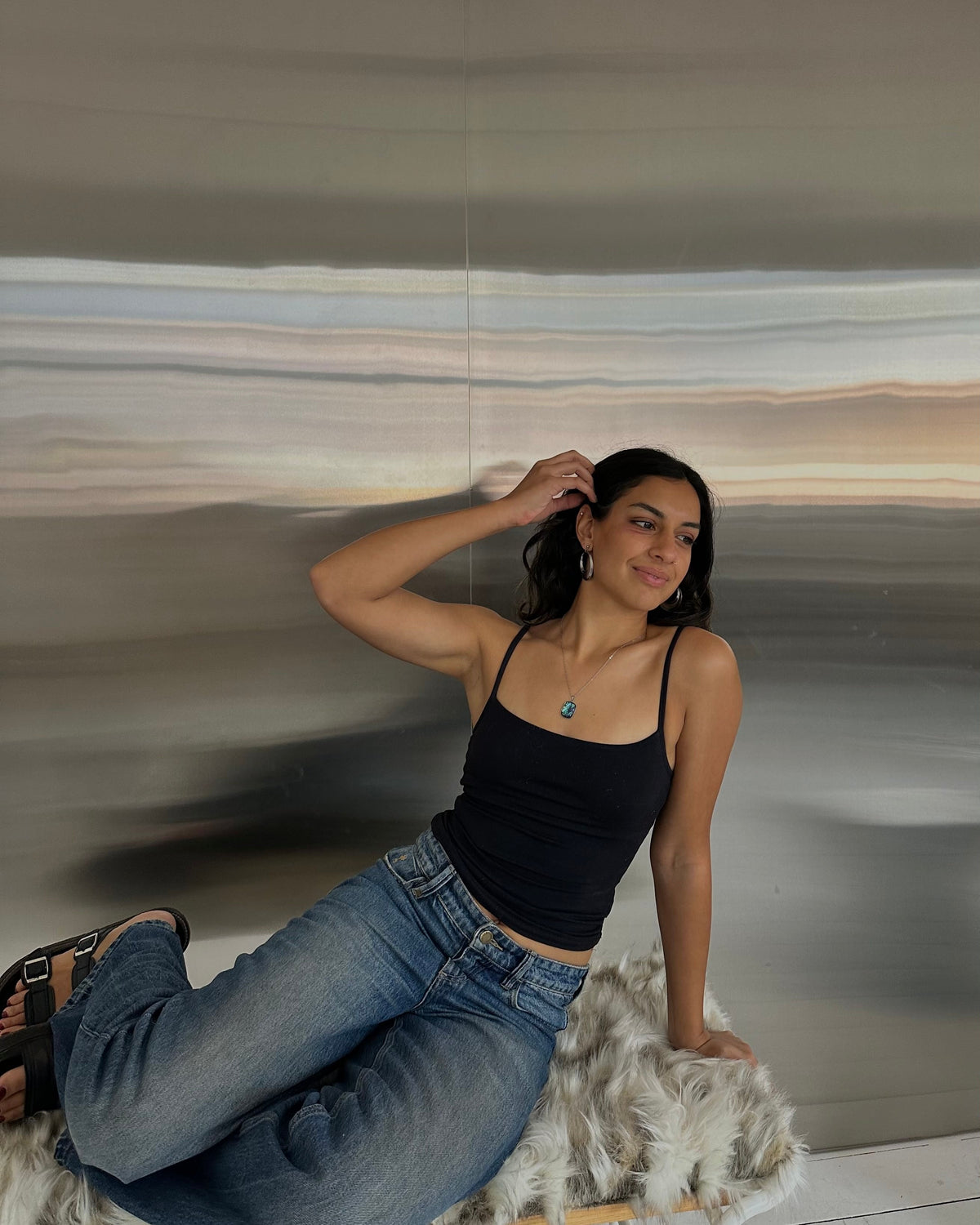 A woman reclining on a white faux-fur bench, wearing a black spaghetti-strap tank top and blue jeans, posed against a large abstract wall art piece.