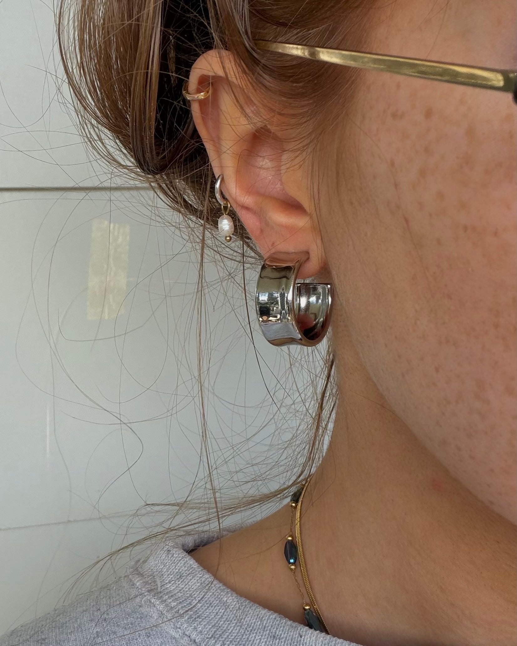 A close-up of a person's ear wearing a large polished silver hoop earring, with other small jewelry pieces also visible against a light indoor background.