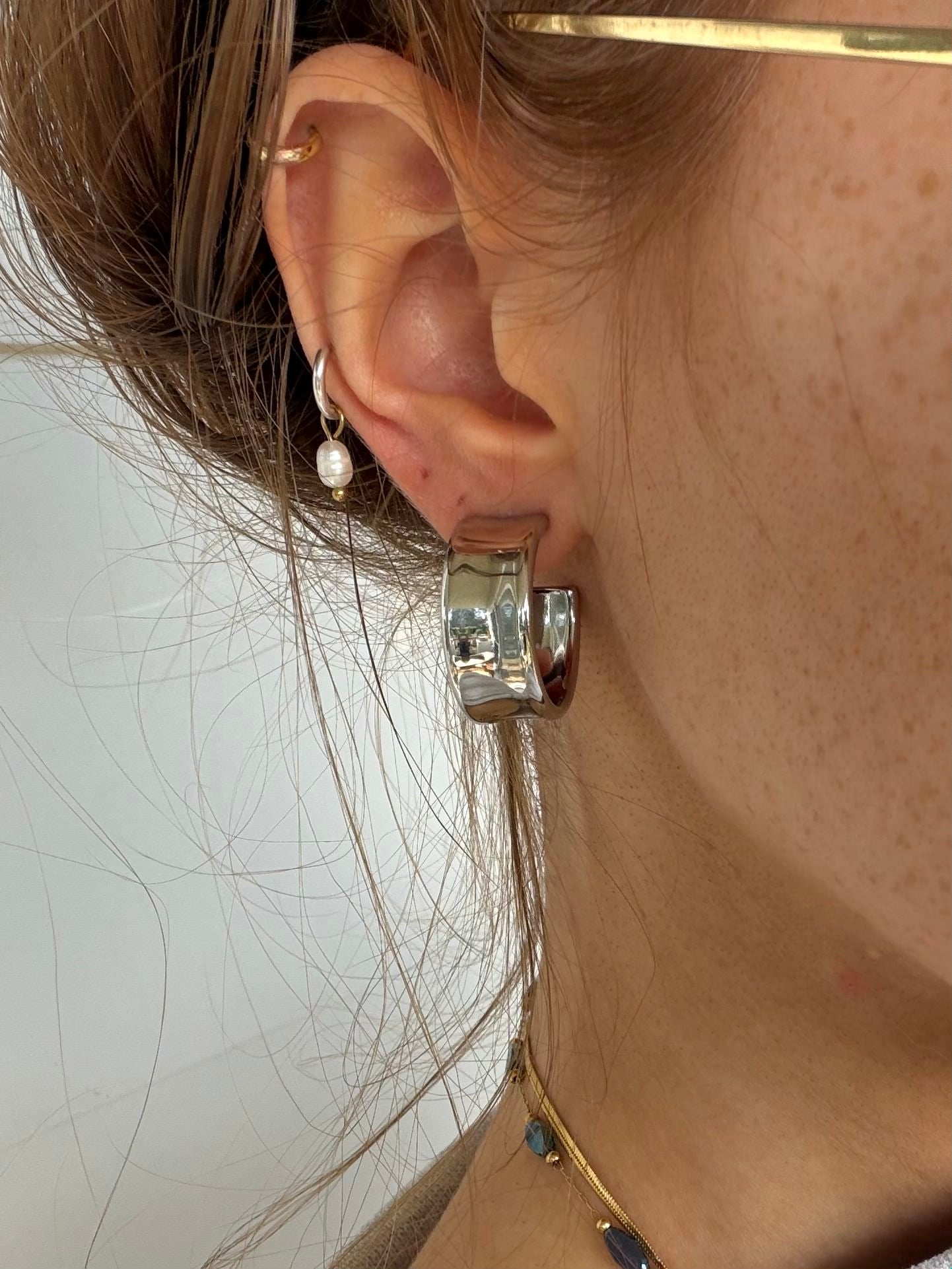 A close-up of a person's ear wearing a chunky silver hoop earring with a polished cylindrical end, along with other small gold and pearl earrings.