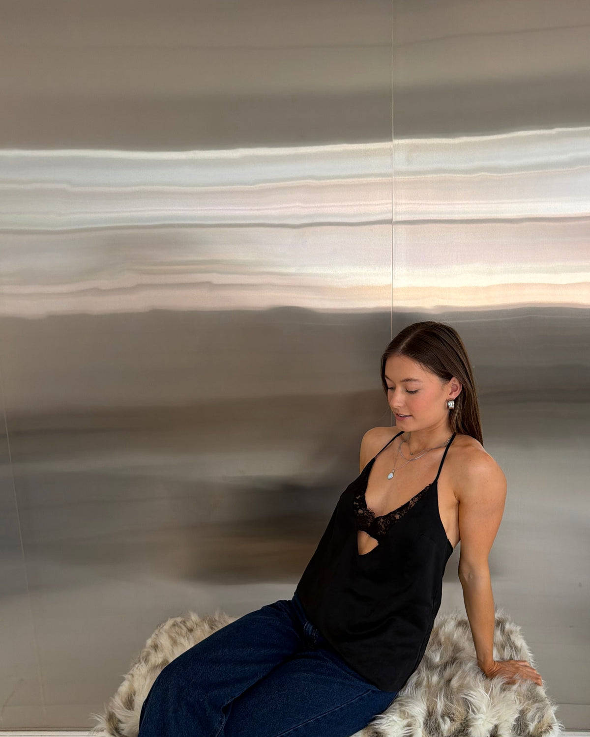 A woman sits on a white bench with a faux-fur throw, wearing a black lace-up camisole top with a chest cutout, dark wide-leg jeans, and gray sneakers, posed against a reflective metallic wall.