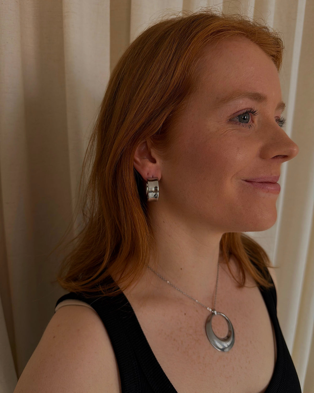 A woman with red hair shown in profile wearing a silver geometric ring pendant necklace and a matching silver stud earring, set against a soft beige curtain background.