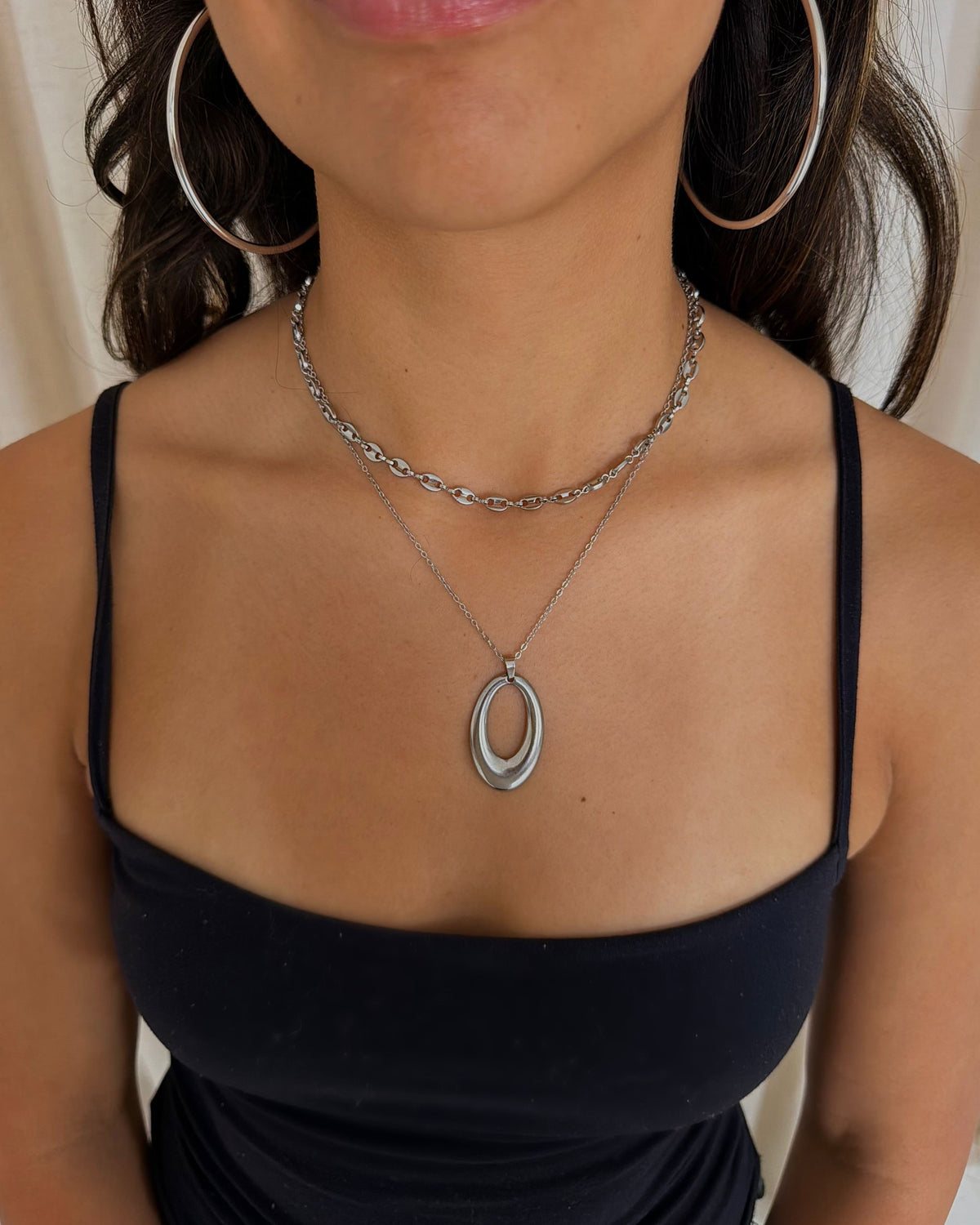 A close-up of a woman wearing layered silver necklaces, including a double oval pendant necklace and a chain necklace, along with large hoop earrings and a black camisole top.