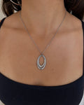 A close-up of a woman wearing a silver double oval pendant necklace, a silver hoop earring, and a black camisole top against a light neutral curtain background.
