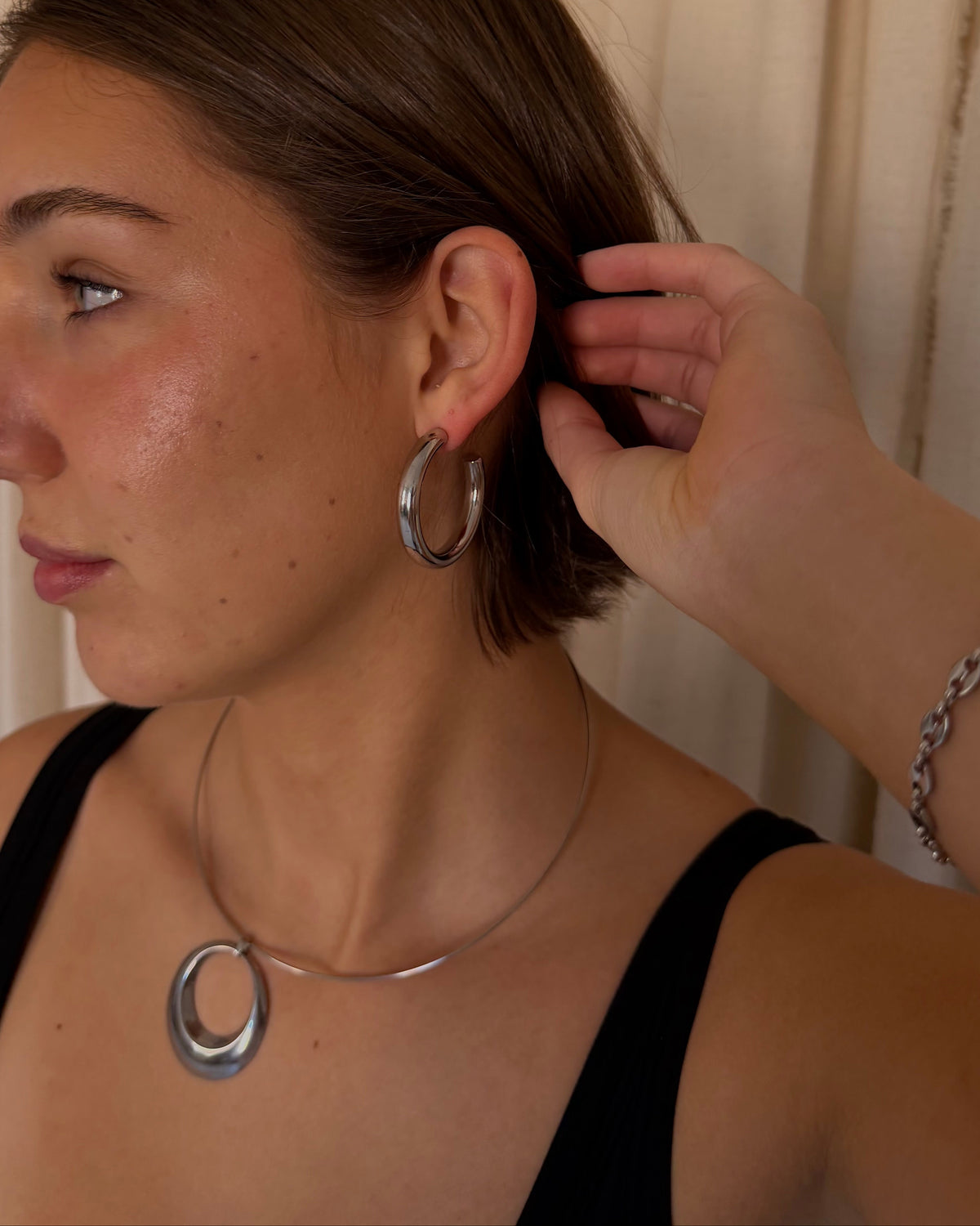 A woman in a black top wearing a silver circular hoop earring and a silver circular pendant necklace, with her hand adjusting the earring. She is standing in front of beige curtains.