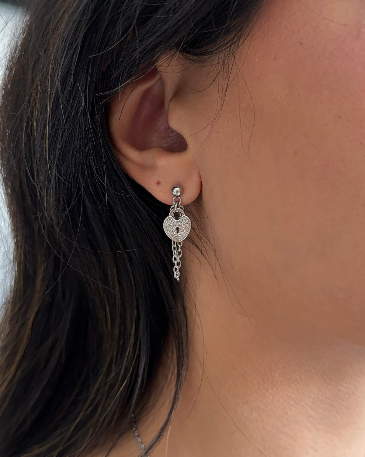 A close-up of a person's ear wearing a silver dangle earring with a heart-shaped charm and a dangling chain.