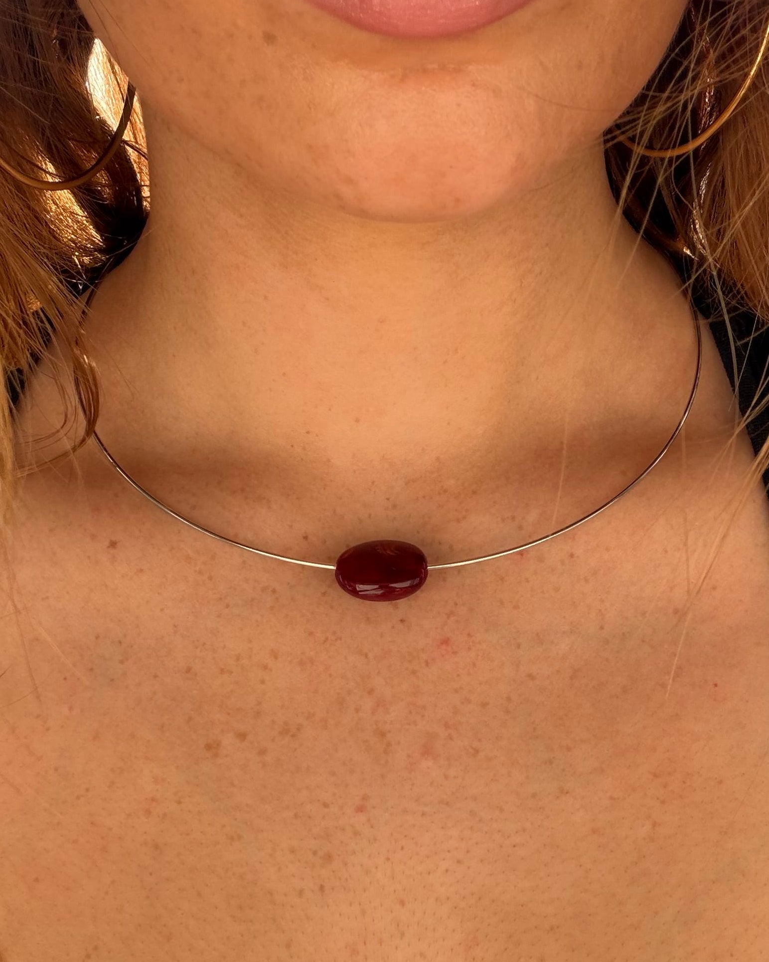 A close-up of a woman's neck and chest wearing a delicate silver wire choker necklace with a single oval dark red gemstone pendant.