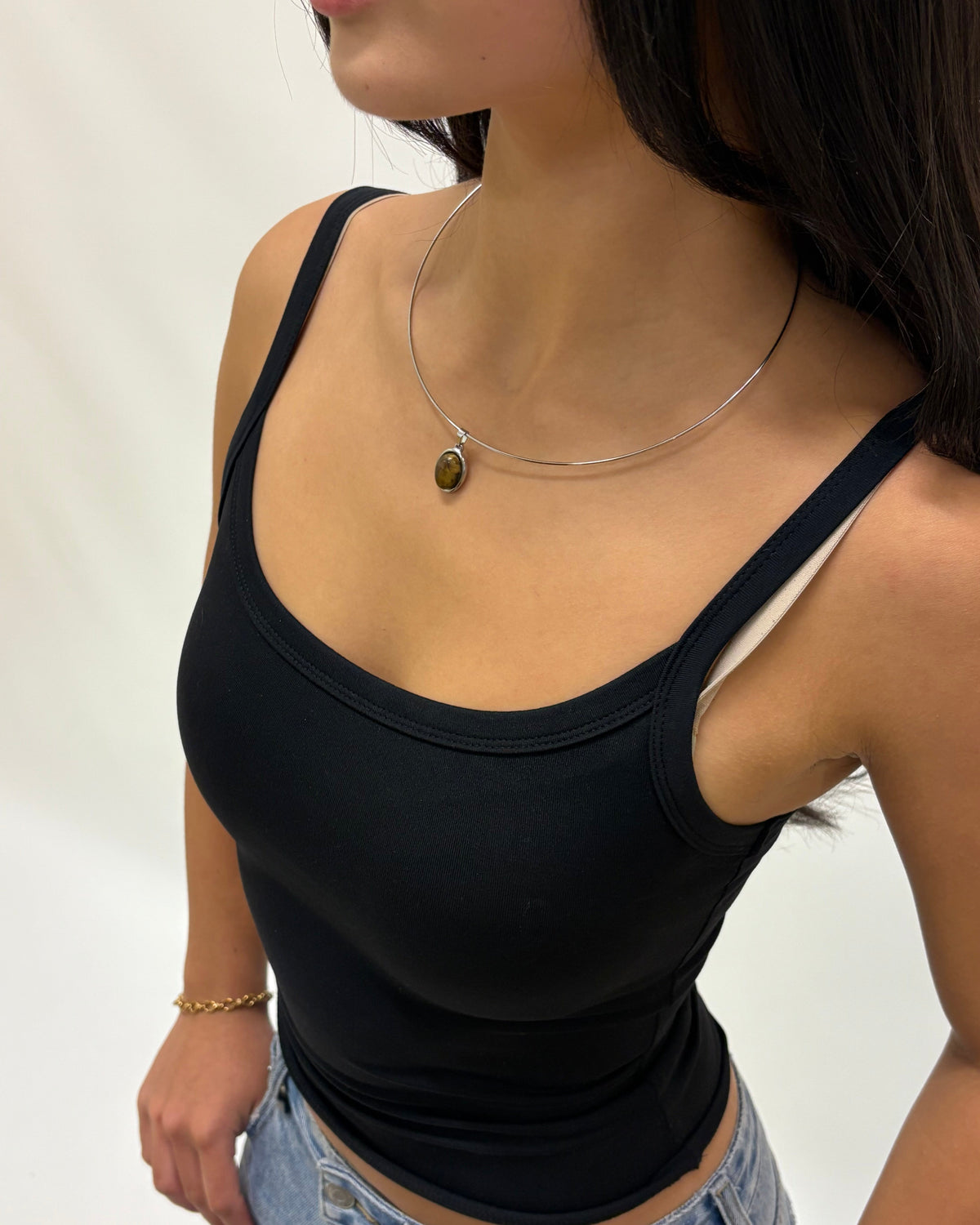 A woman wearing a fitted black ribbed tank top with thin straps and a scoop neckline, paired with light blue jeans. She is also wearing a delicate silver necklace with a small oval pendant.
