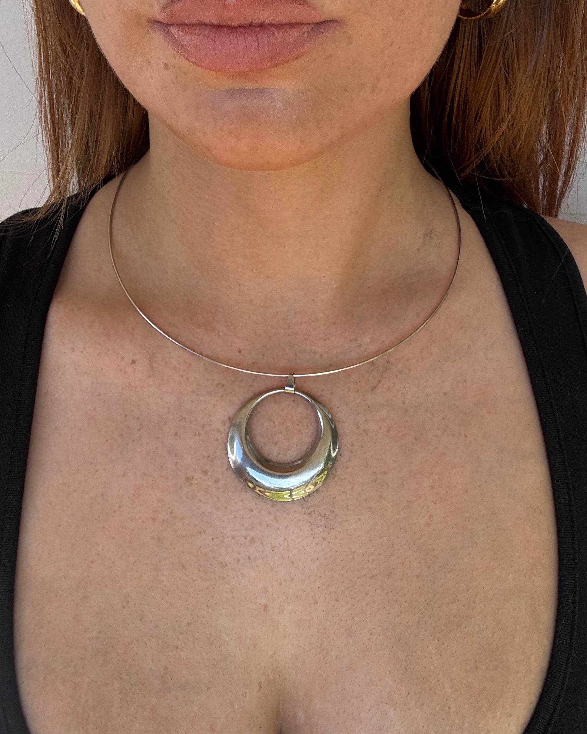 A close-up of a person lying down wearing a silver circular ring collar necklace and a thin silver wire collar, with a black sports bra and gold hoop earrings visible.
