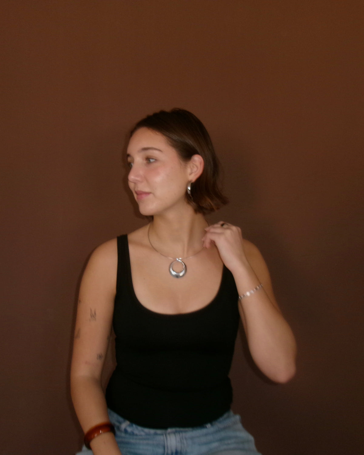 A woman with short brown hair wearing a black tank top and light blue jeans, wearing a silver circular pendant necklace and hoop earrings, posing against a solid dark brown background.