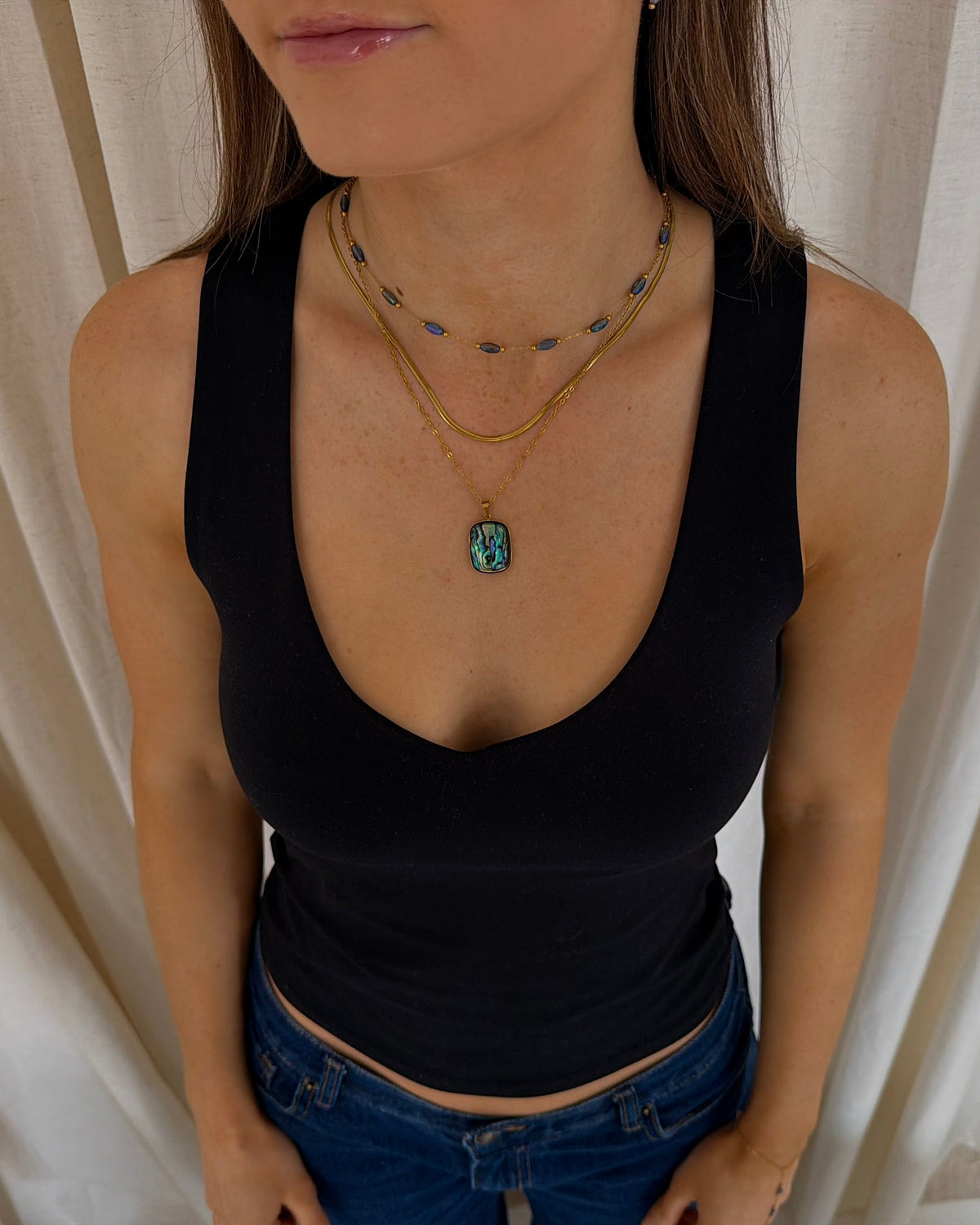 A woman wearing a black tank top and blue jeans, showcasing a layered gold necklace set with a turquoise-colored pendant against a neutral curtain background.