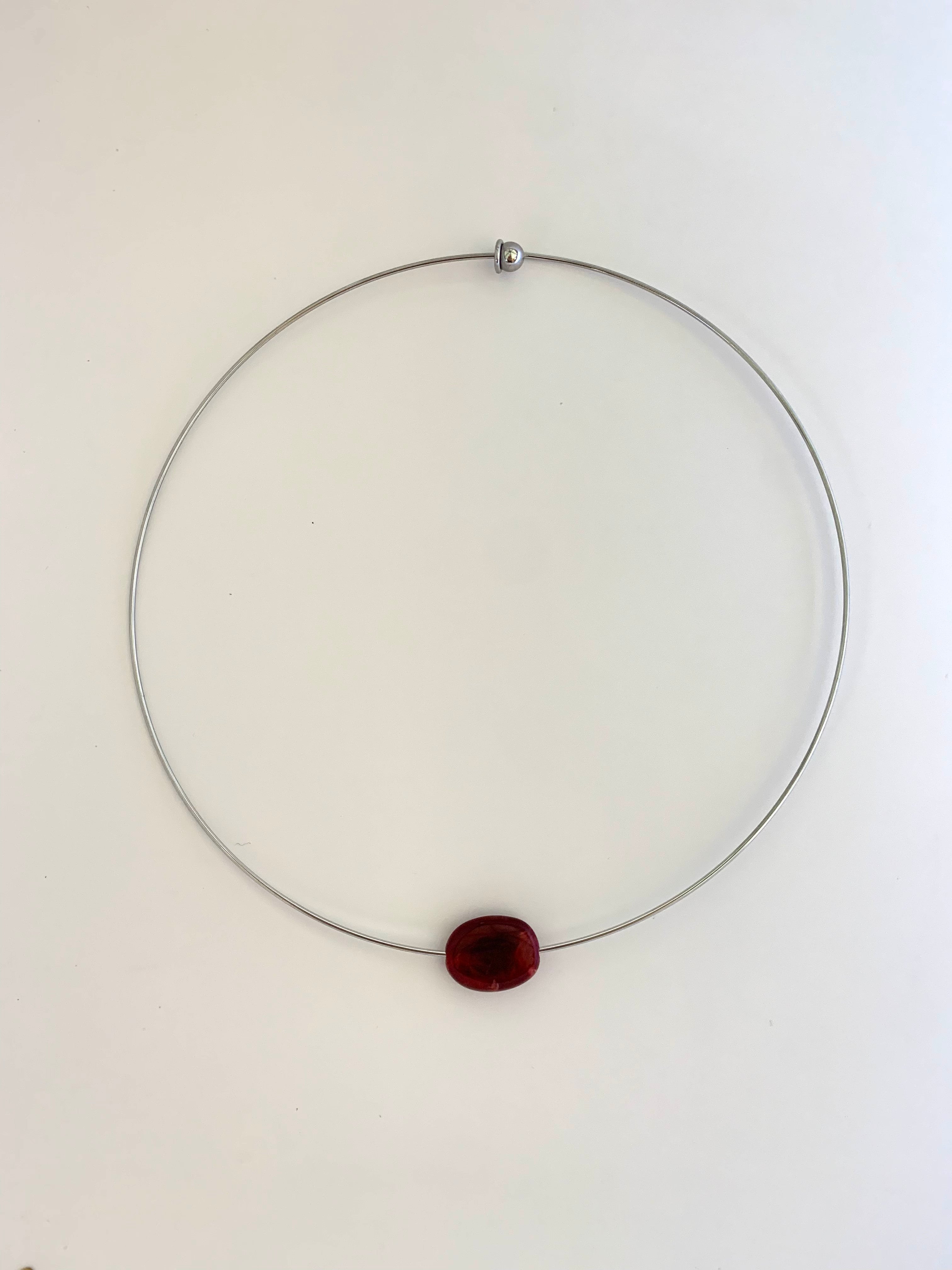 A minimalist silver wire necklace with a single small, oval, dark red bead pendant, displayed against a plain white background.