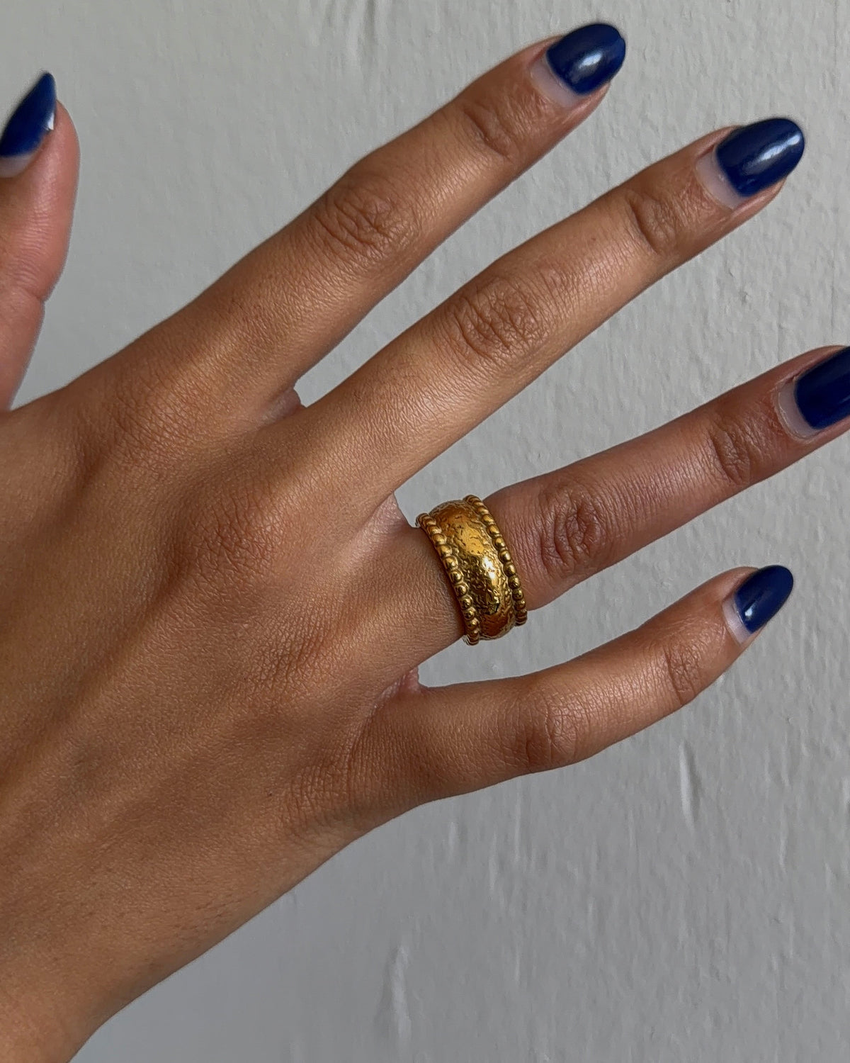 A close-up of a hand with dark blue nail polish wearing a textured gold ring, set against a neutral light gray wall.