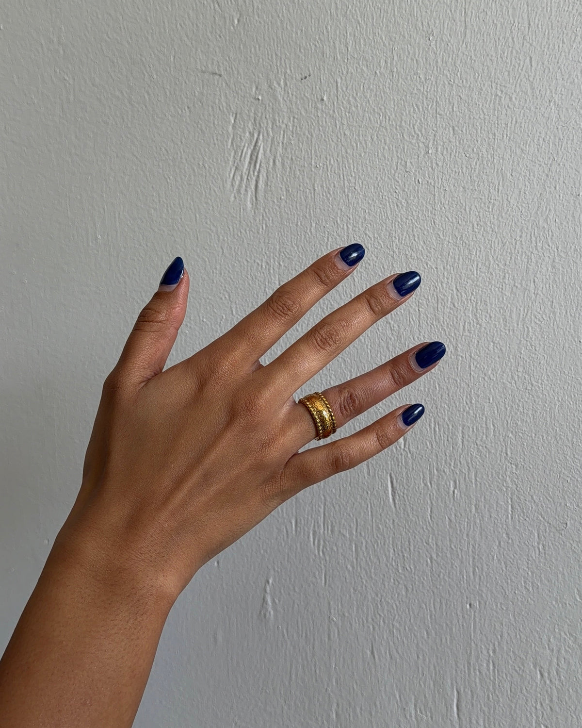 A hand with dark blue nail polish wearing gold rings, set against a textured off-white wall.