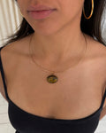 A close-up of a model wearing a delicate gold wire necklace with an oval pendant containing a yellow-green and black pattern, tigers eye stone paired with gold hoop earrings and a black top.