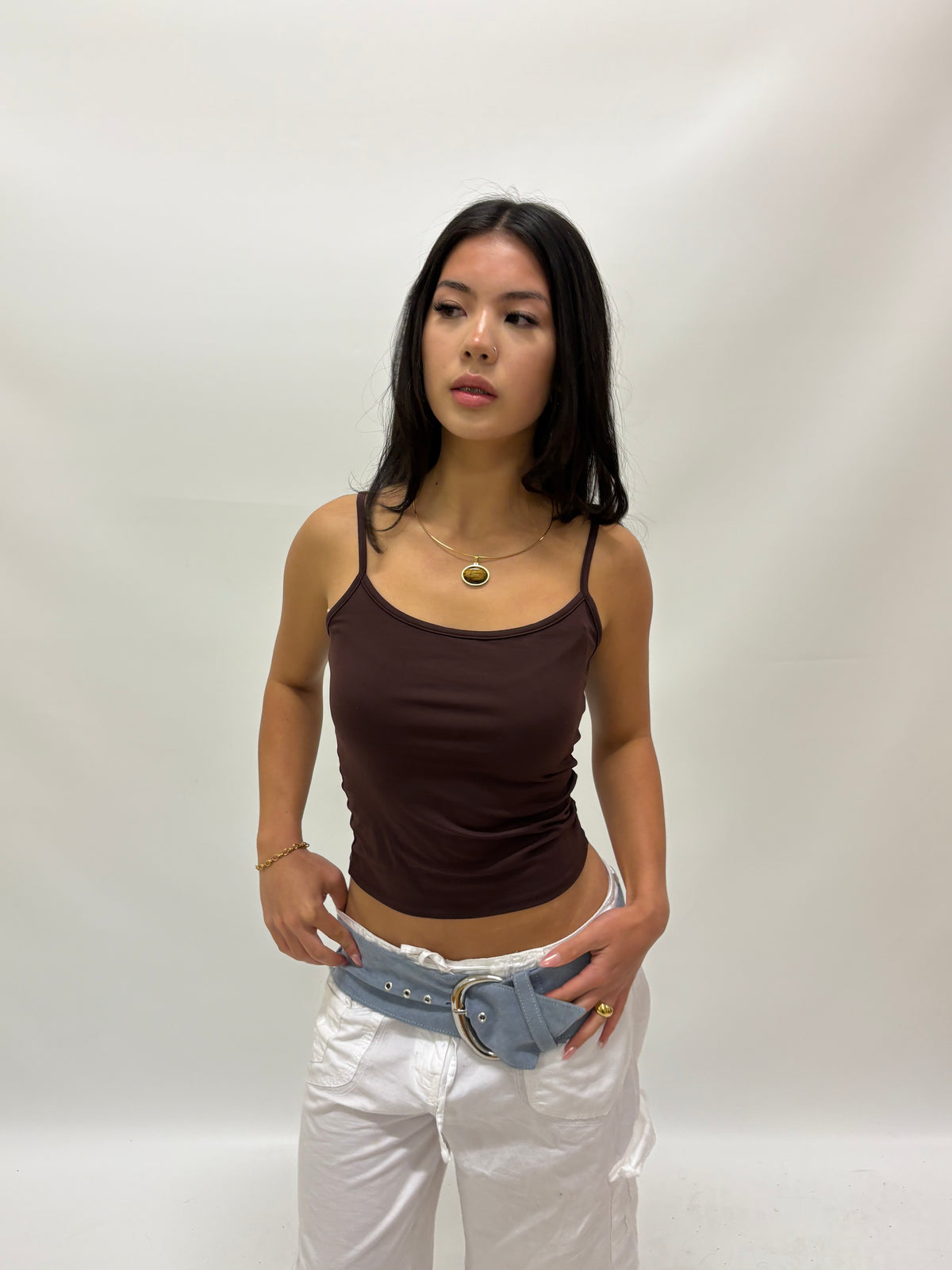 A close-up of a model wearing a delicate gold wire necklace with an oval pendant containing a yellow-green and black pattern, tigers eye stone paired with gold hoop earrings and a brown top.