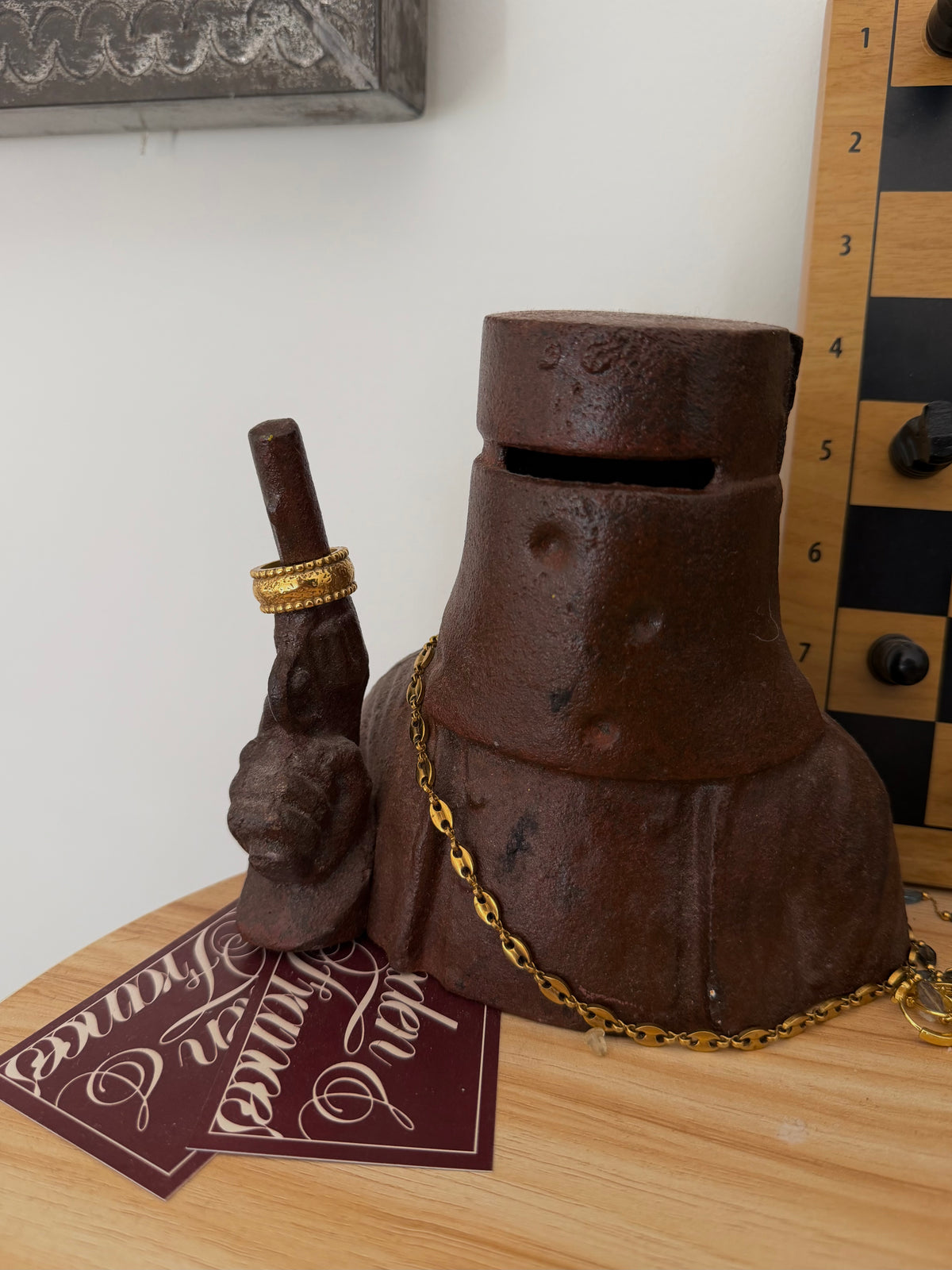 A rustic dark brown metal bell-shaped container with a coin slot, styled like an old knight's helmet or shield, with a gold chain draped around it. It is shown on a wooden tabletop next to a gold ring, a card, and a checkered board in the background.