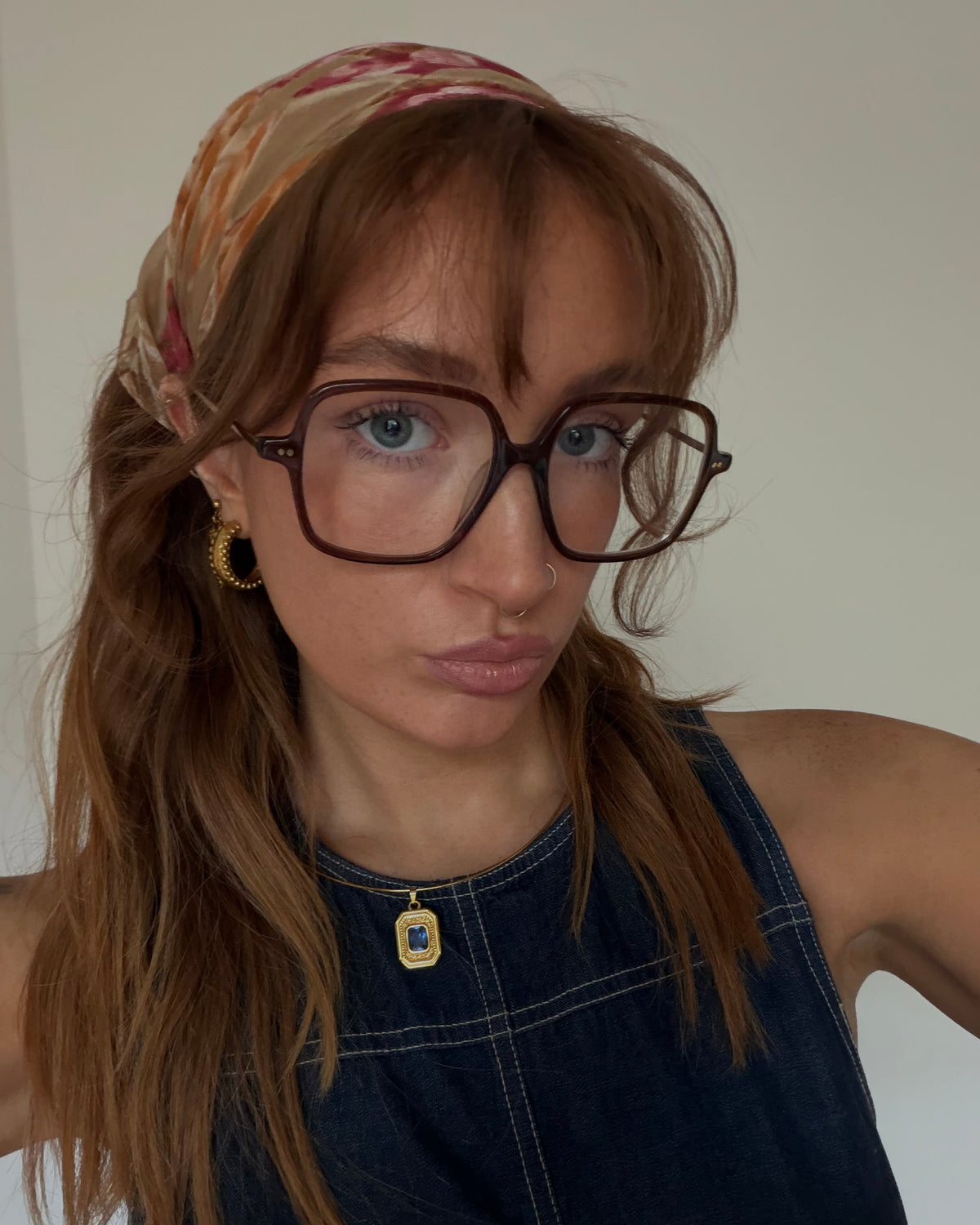 A woman with long light brown hair wearing a patterned headscarf, thick brown-rimmed eyeglasses, a gold pendant necklacewith a blue stone and textured gold hoop earrings