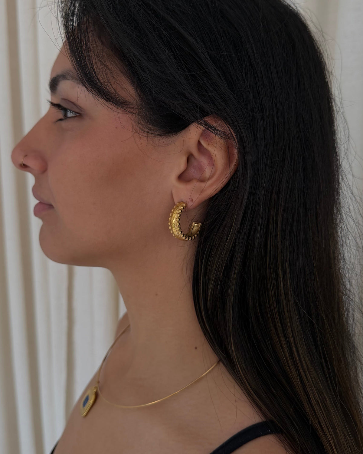 A woman shown in profile wearing a textured beaded gold hoop earrings and a wire necklace