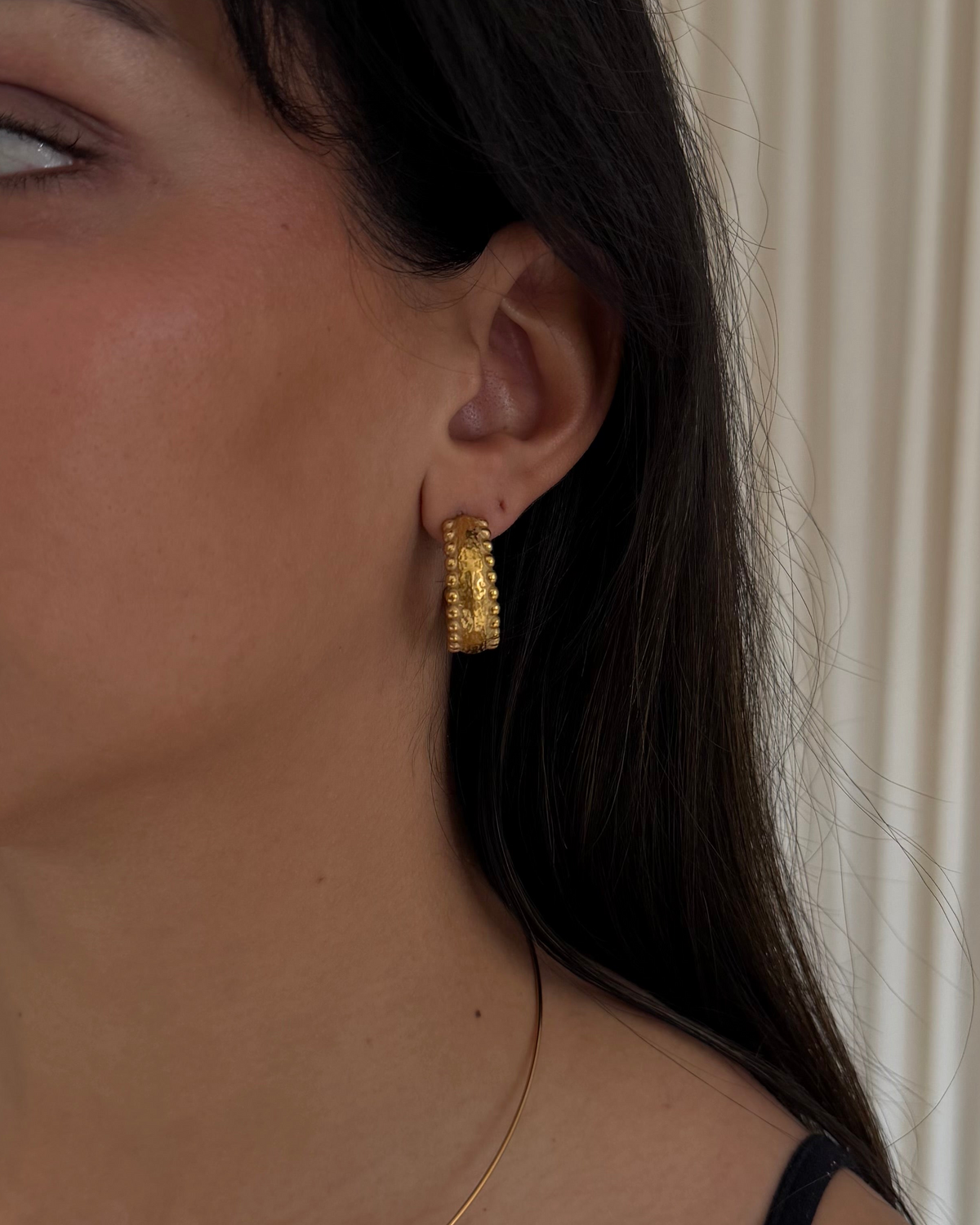 A close-up of a woman's ear wearing a textured gold hoop earring, with beading along the sides.