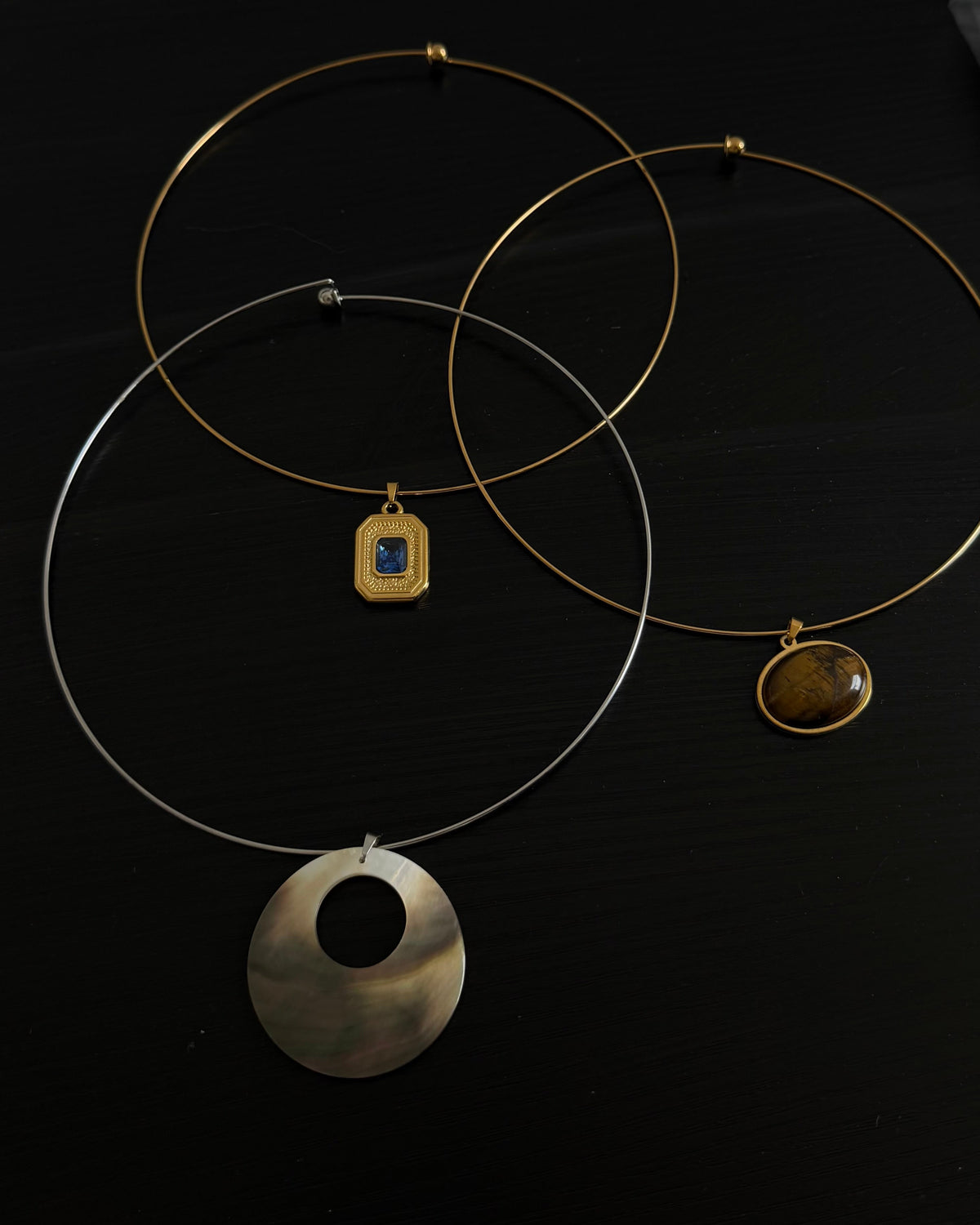 Three minimalist wire necklaces with circular and rectangular pendants in gold and silver tones, displayed on a dark surface.