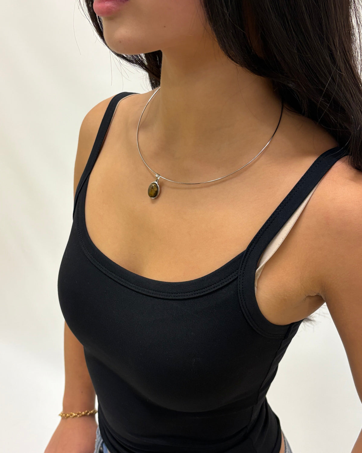 A close-up of a woman wearing a thin silver wire necklace with a small oval brown tiger's eye pendant, paired with a black tank top.