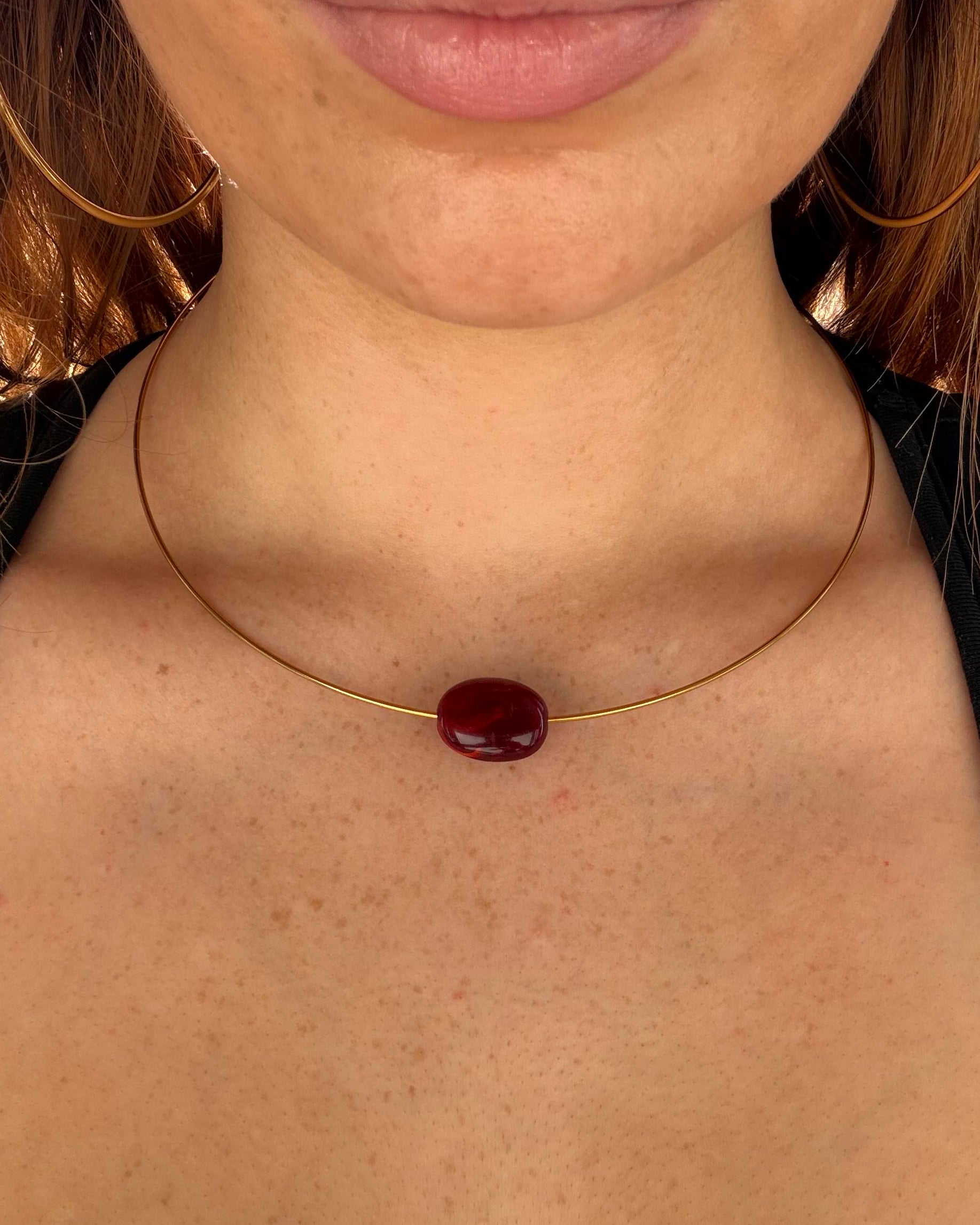 A close-up of a person wearing a delicate gold necklace with a single oval dark red gemstone pendant, styled with large gold hoop earrings.