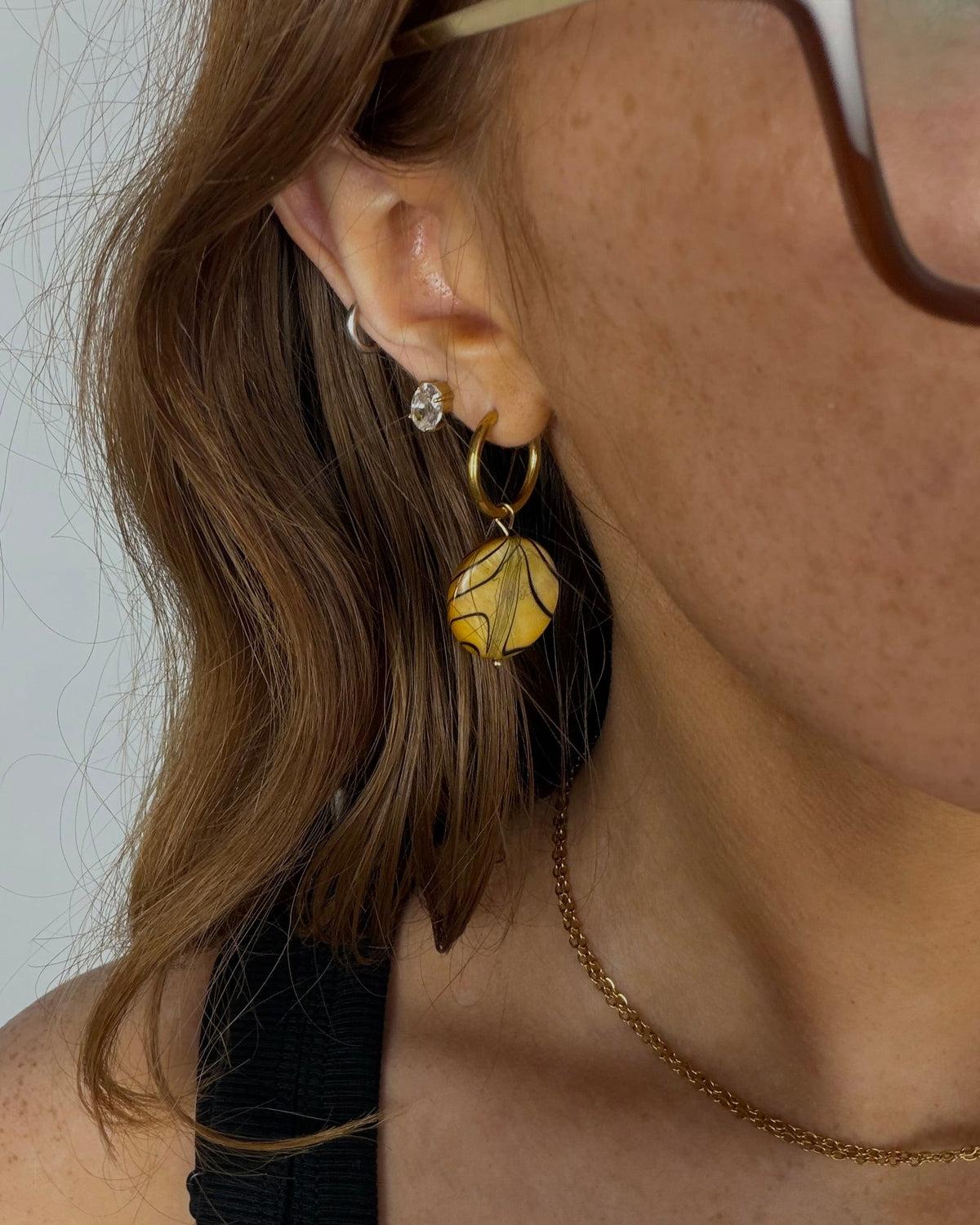 A close-up of a person's ear wearing a gold hoop earring with a textured yellow-green leaf dangle, alongside other small earrings and glasses.