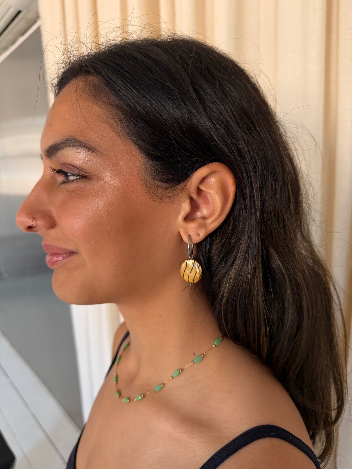 A woman shown in profile wearing a tiger print earring and a matching green and gold beaded necklace, set against a soft beige curtain background.