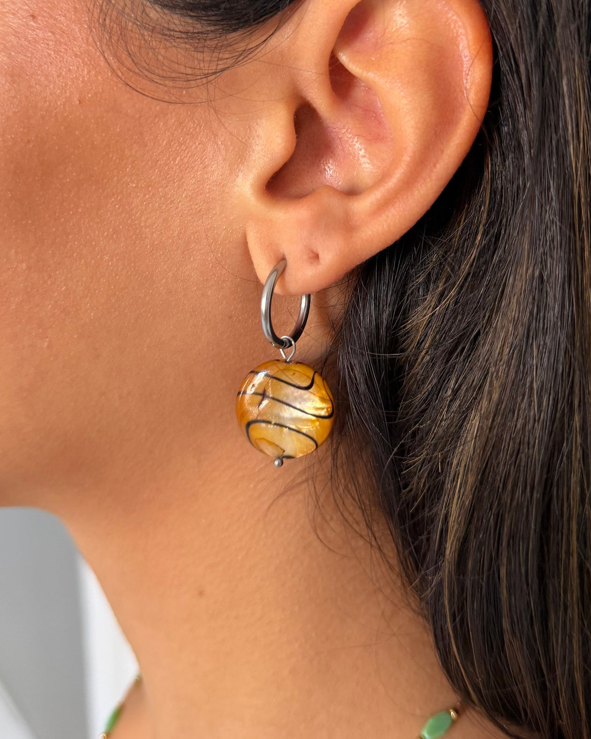 Close-up of a woman's ear wearing a silver huggie hoop earring with a dangling marbled yellow, gold, and brown glass drop earring.