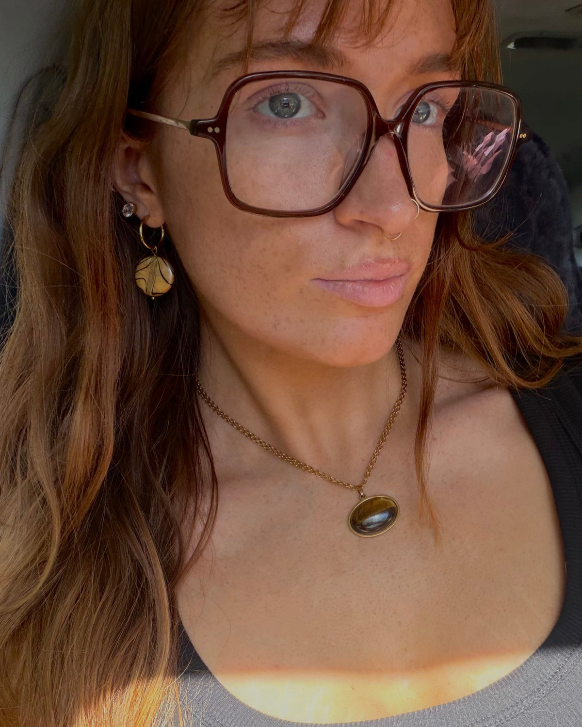 A woman with long light brown hair, wearing large brown-rimmed square glasses, a black tank top, and a gold pendant necklace, sitting inside a vehicle.