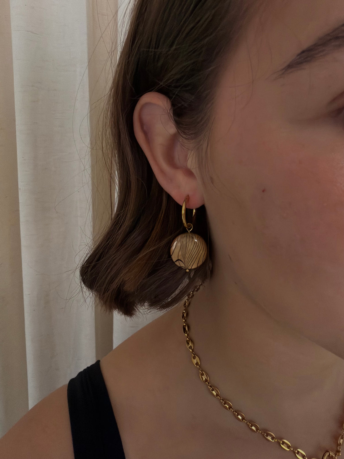 A close-up of a person wearing a gold huggie hoop earring with a textured, hammered circular disc pendant. The person has brown hair and is wearing a black top and a gold chain necklace, set against a soft beige curtain background.