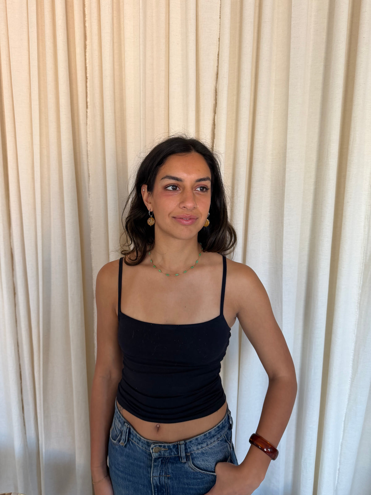 A woman wearing a black spaghetti-strap crop top and blue jeans, standing in front of a cream-colored pleated curtain backdrop. vintage colourful jewellery waterproof