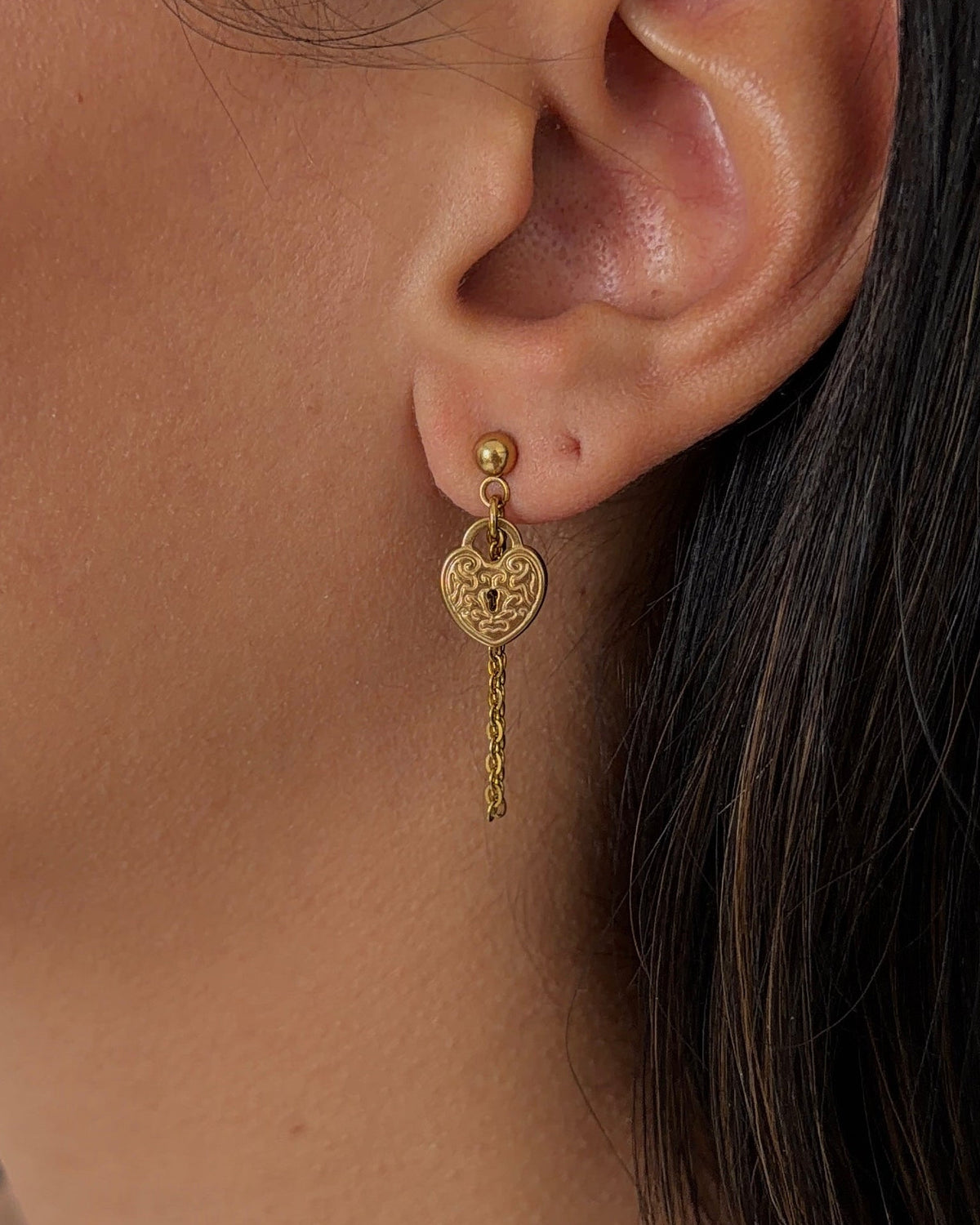 A close-up of a person's ear wearing a gold dangle earring with a heart-shaped charm and a dangling chain.